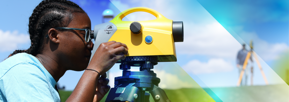 Woman at bottom of grassy hill, looking into surveying equipment. Soft focus background on another student